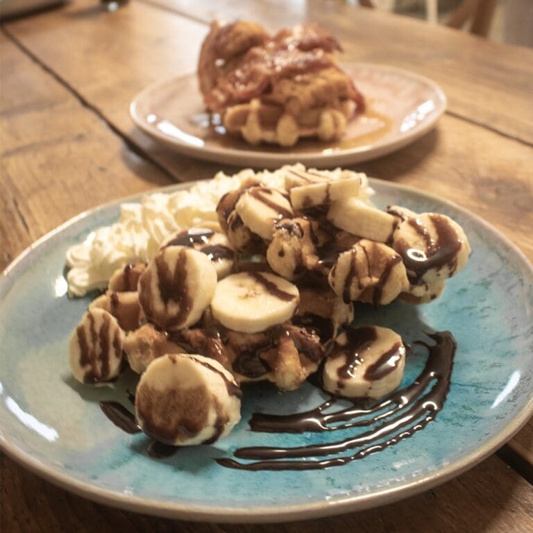 This is a waffle topped with sliced banana, chocolate sauce and whipped cream.